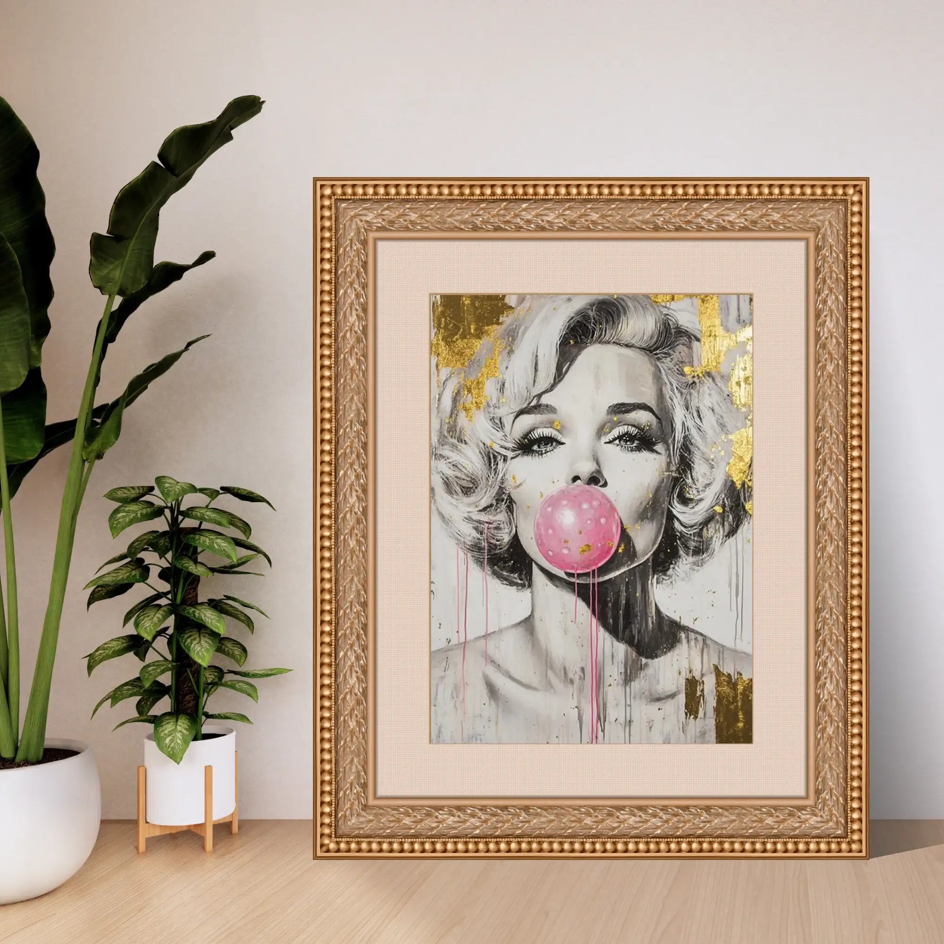 Framed artwork of a stylized portrait with a pink bubble gum bubble, placed on a light wooden floor with plants in the background.