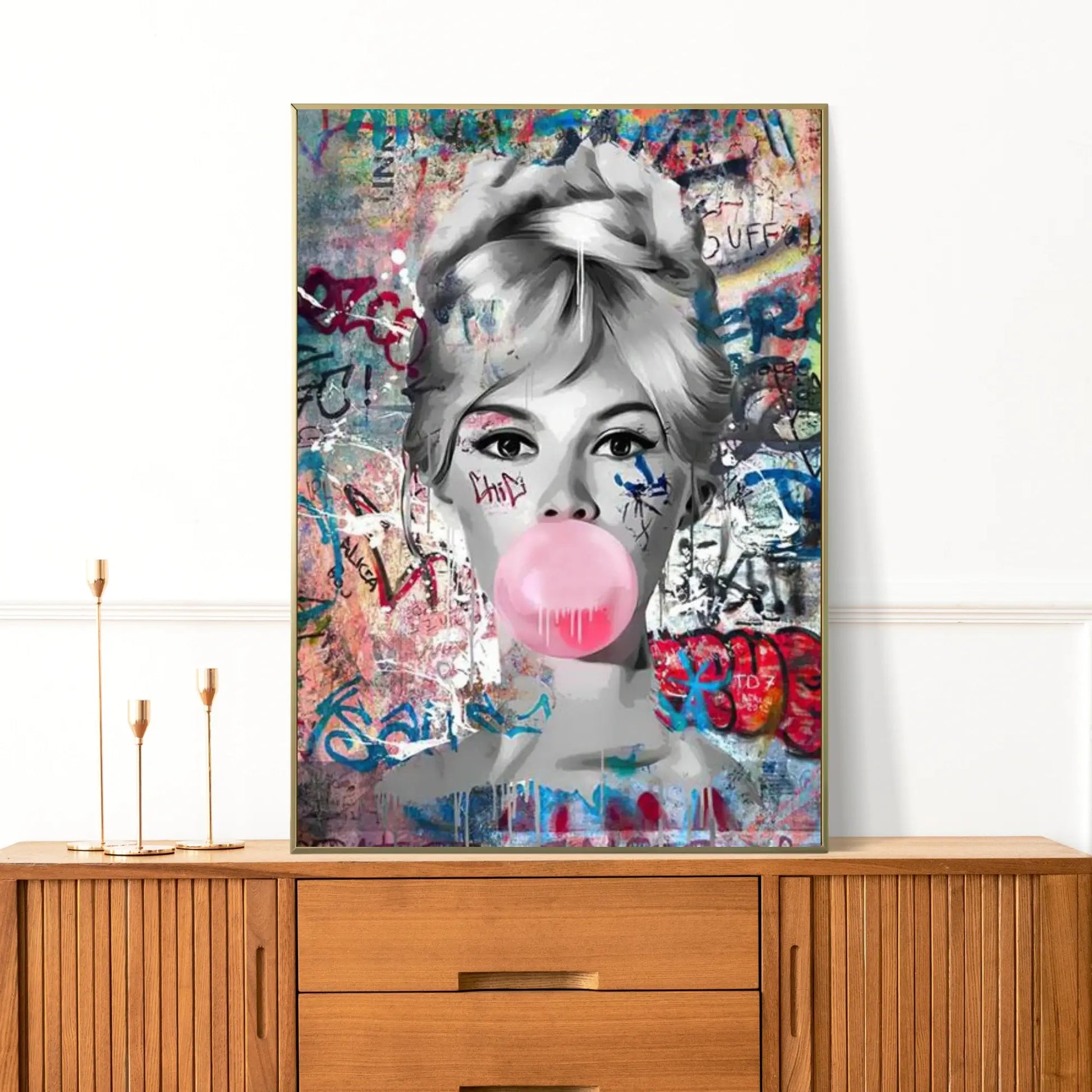 Framed artwork of a woman blowing a pink bubble gum bubble on a colorful background, displayed on a wooden cabinet.